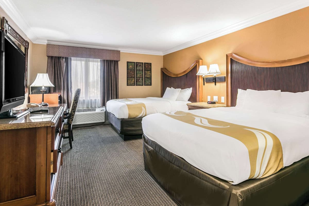 Quarto de hotel com duas camas, uma mesa de trabalho, televisão e janela com cortinas.