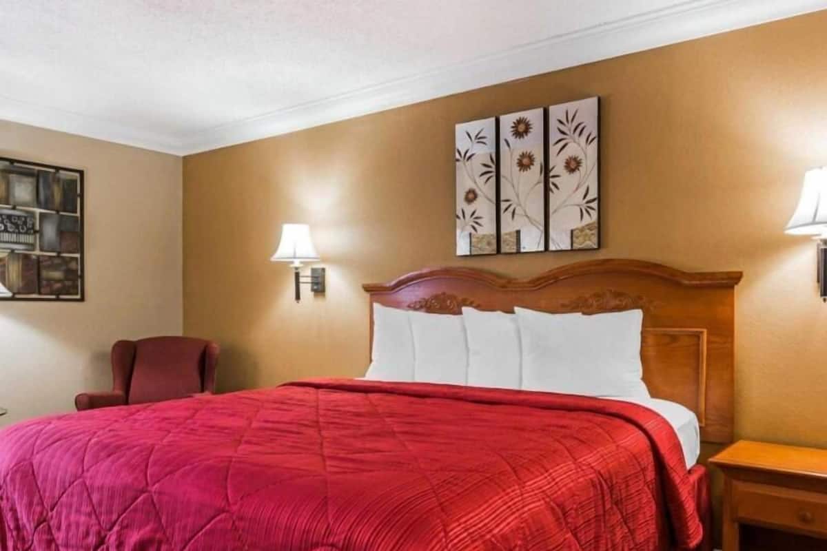 Quarto de hotel com uma cama grande, cabeceira de madeira, colcha acolchoada vermelha, luminária fixada na parede e uma obra de arte floral emoldurada sobre a cama.
