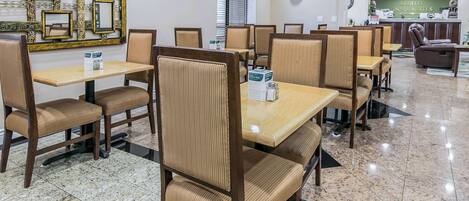A hotel dining area with wooden tables and cushioned chairs, a reception desk, and a decorative wall panel.