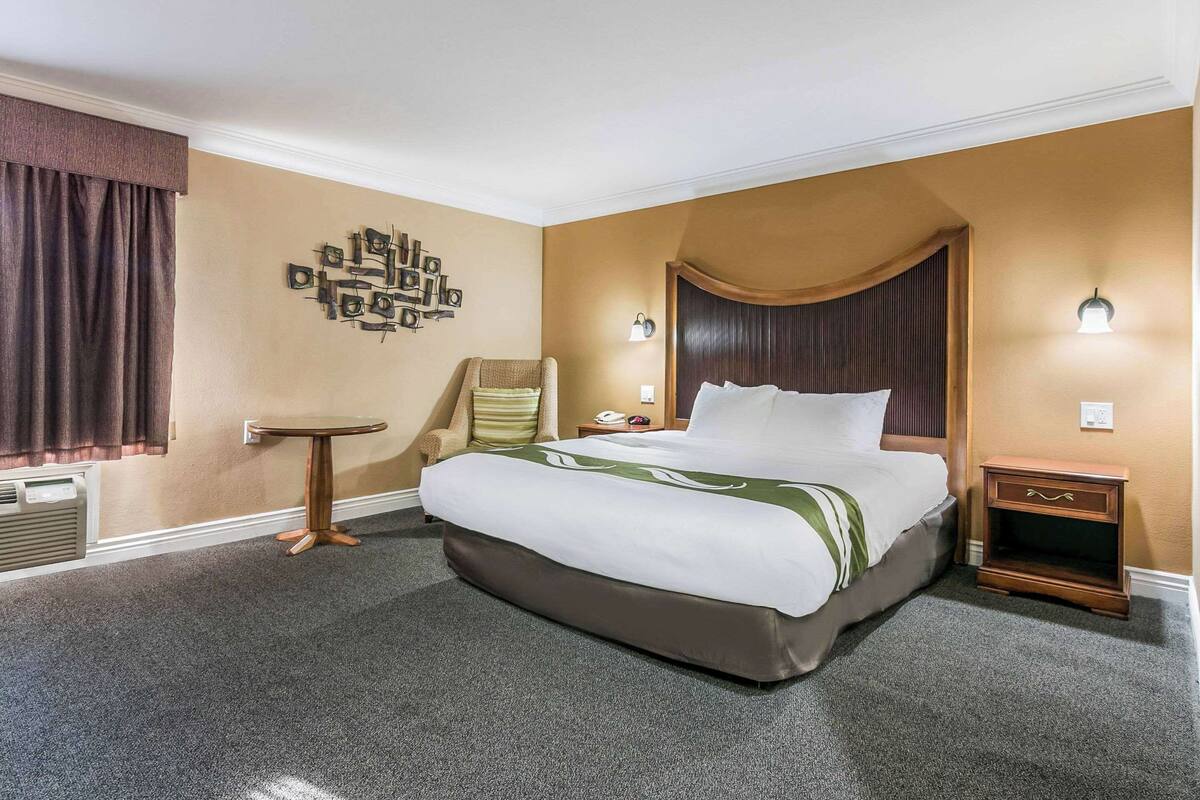 Quarto de hotel com uma cama grande, uma mesinha redonda, uma cadeira, um criado-mudo e uma obra de arte na parede.