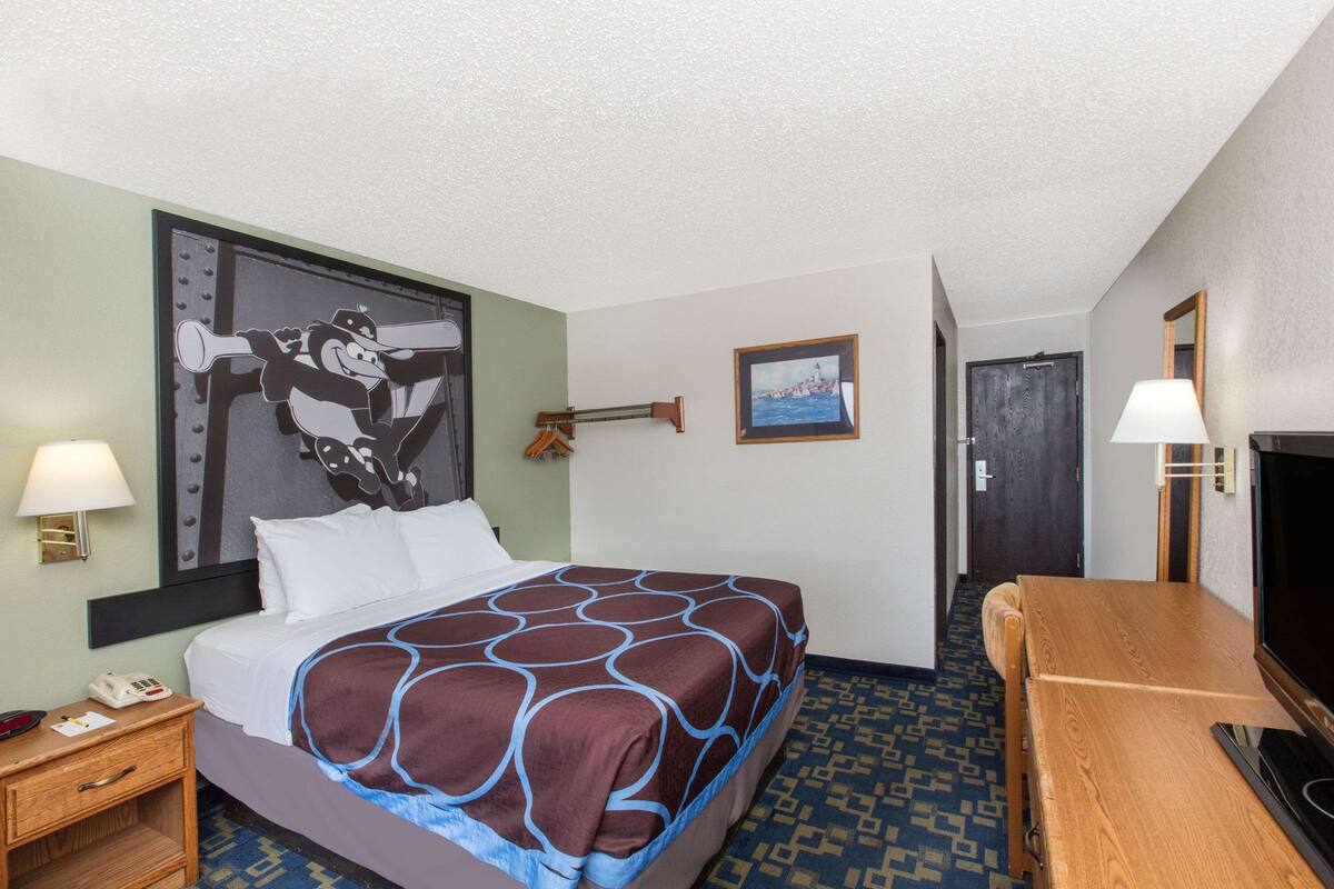 A hotel room with a bed, a desk, a TV, a lamp, a picture frame, and a wall clock.