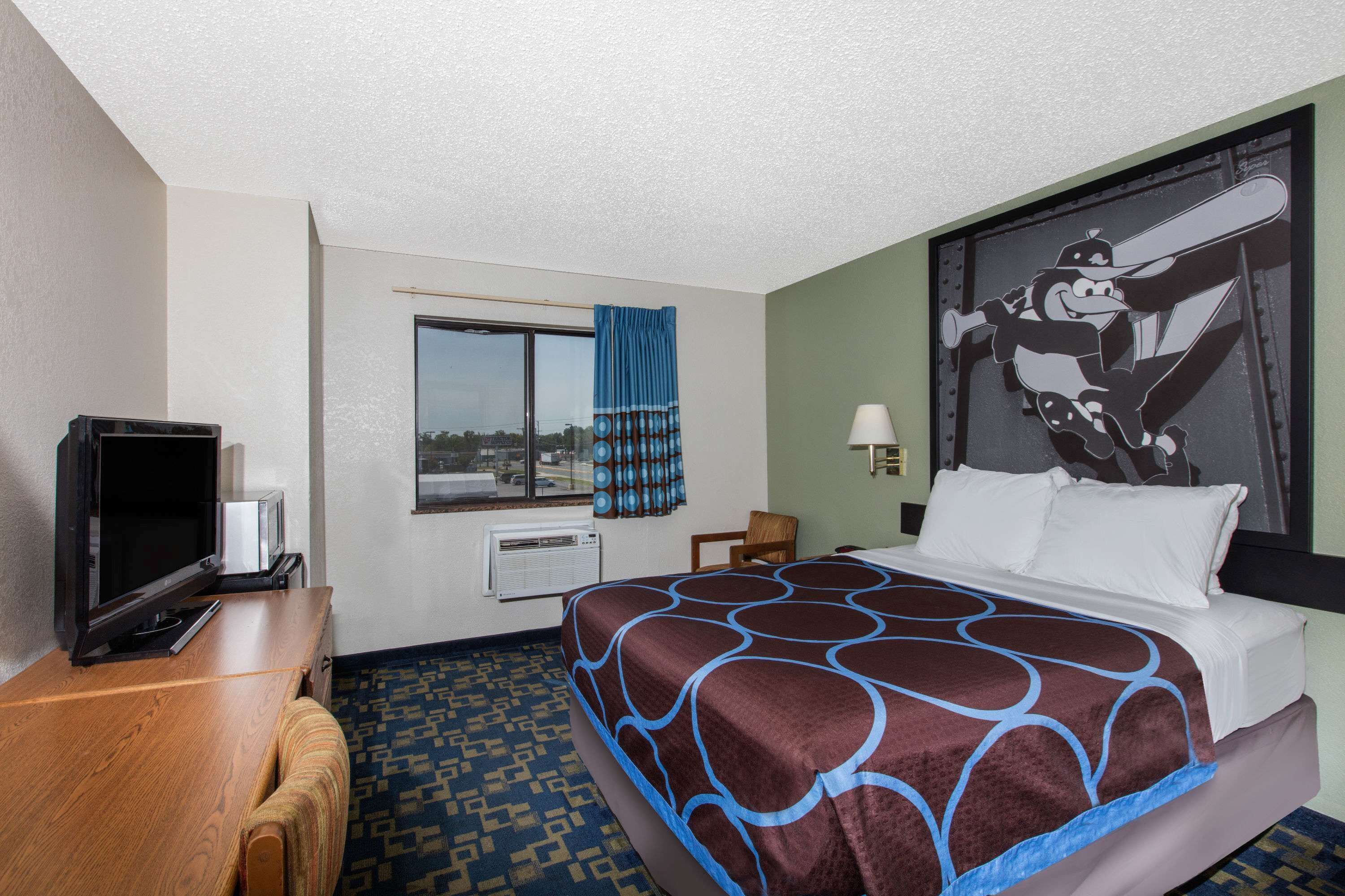 Habitación de hotel con cama, escritorio con televisor, ventana con cortinas y lámpara de pared.