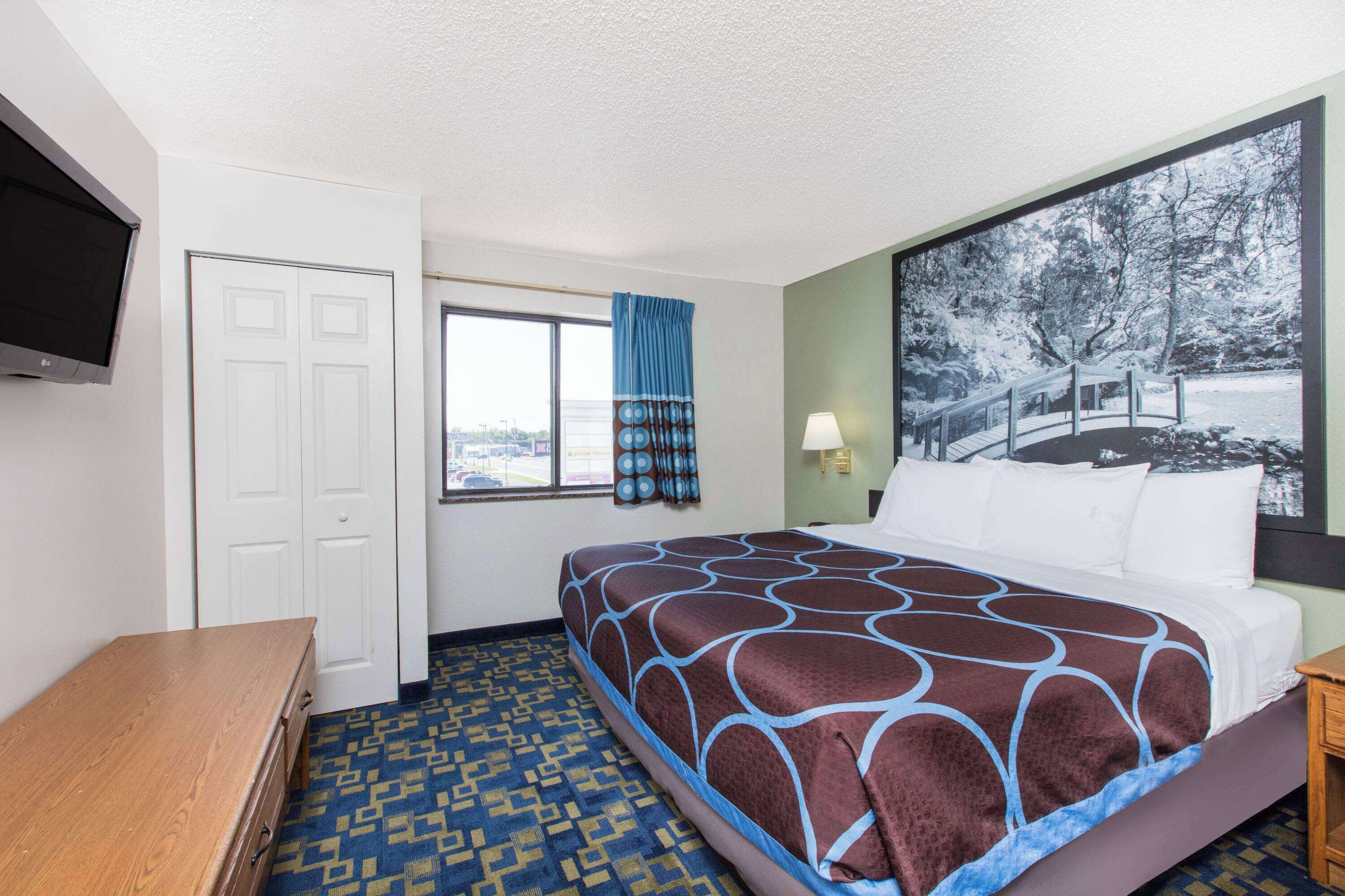 Una habitación de hotel con una cama grande, un aparador de madera, un televisor montado en la pared, una ventana con cortinas y un cuadro de un puente.