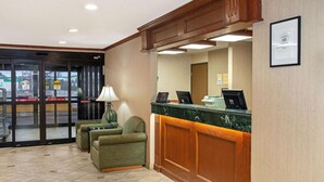 Ein Hotel-Rezeptionsbereich mit einem Holzbüro, zwei grünen Sesseln, einer Lampe und einem gerahmten Zertifikat an der Wand.