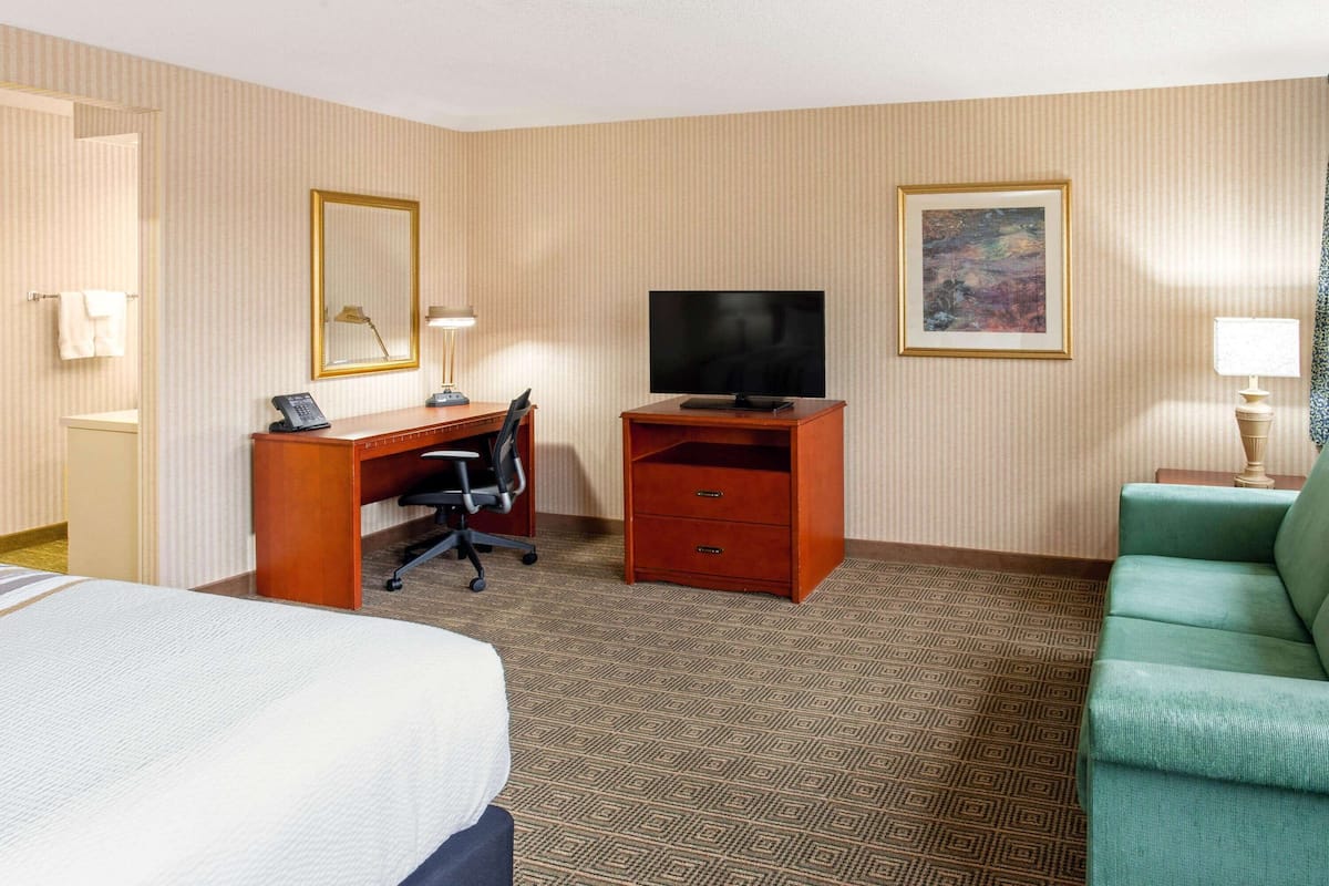 A hotel room with a bed, a desk with a computer, a television, a green sofa, and a patterned carpet.