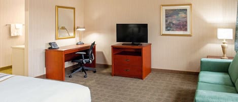 Une chambre d’hôtel avec un lit, un bureau équipé d’un ordinateur, une télévision, un canapé vert et une moquette à motifs.