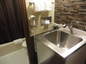 A bathroom with a stainless steel sink, a hair dryer on the wall, and a tiled floor.