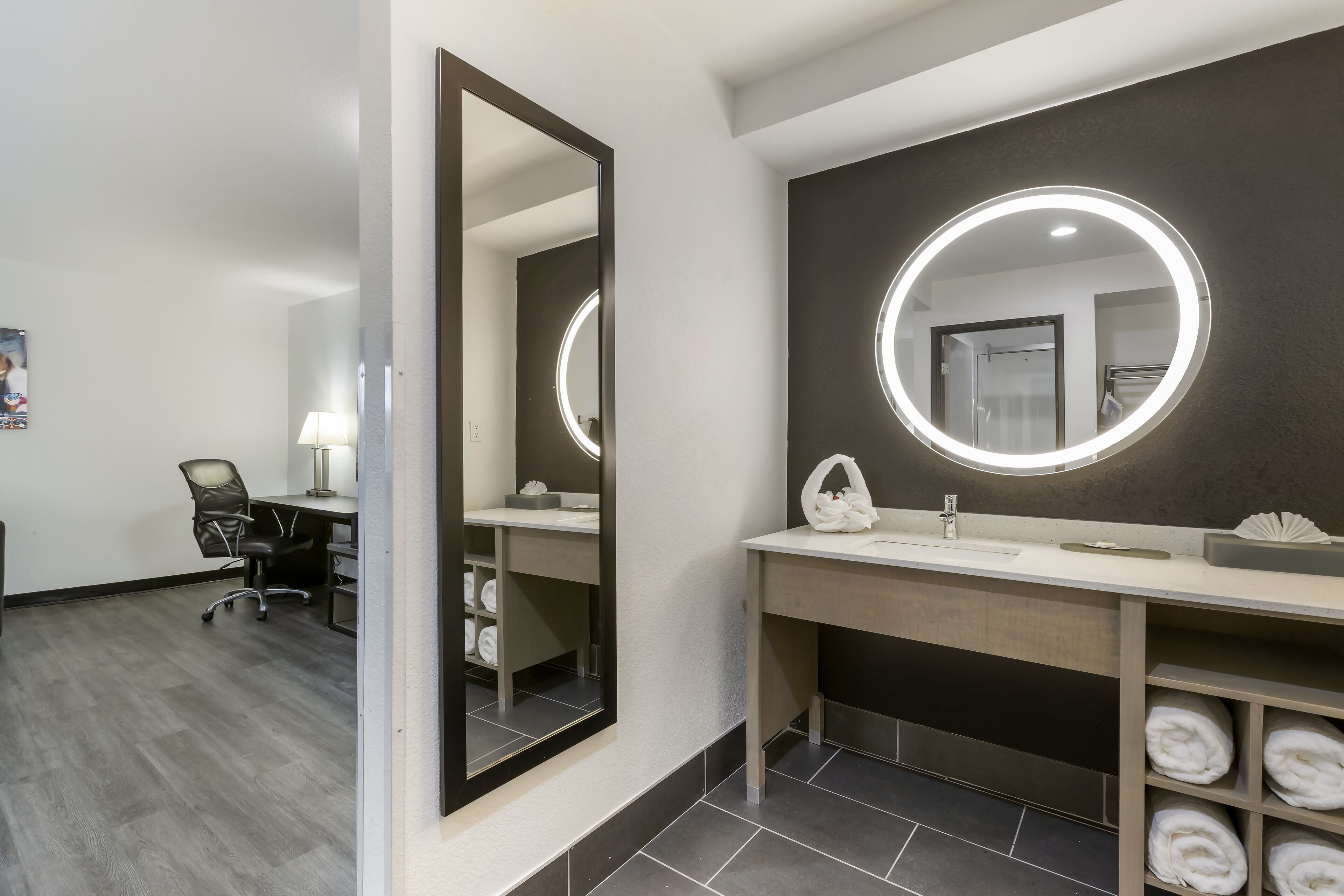Un bagno moderno con un ampio specchio, uno specchio rotondo illuminato e un mobile lavabo con asciugamani.