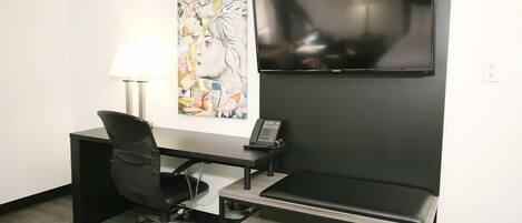 A hotel room with a desk, chair, TV, artwork, and a lamp.