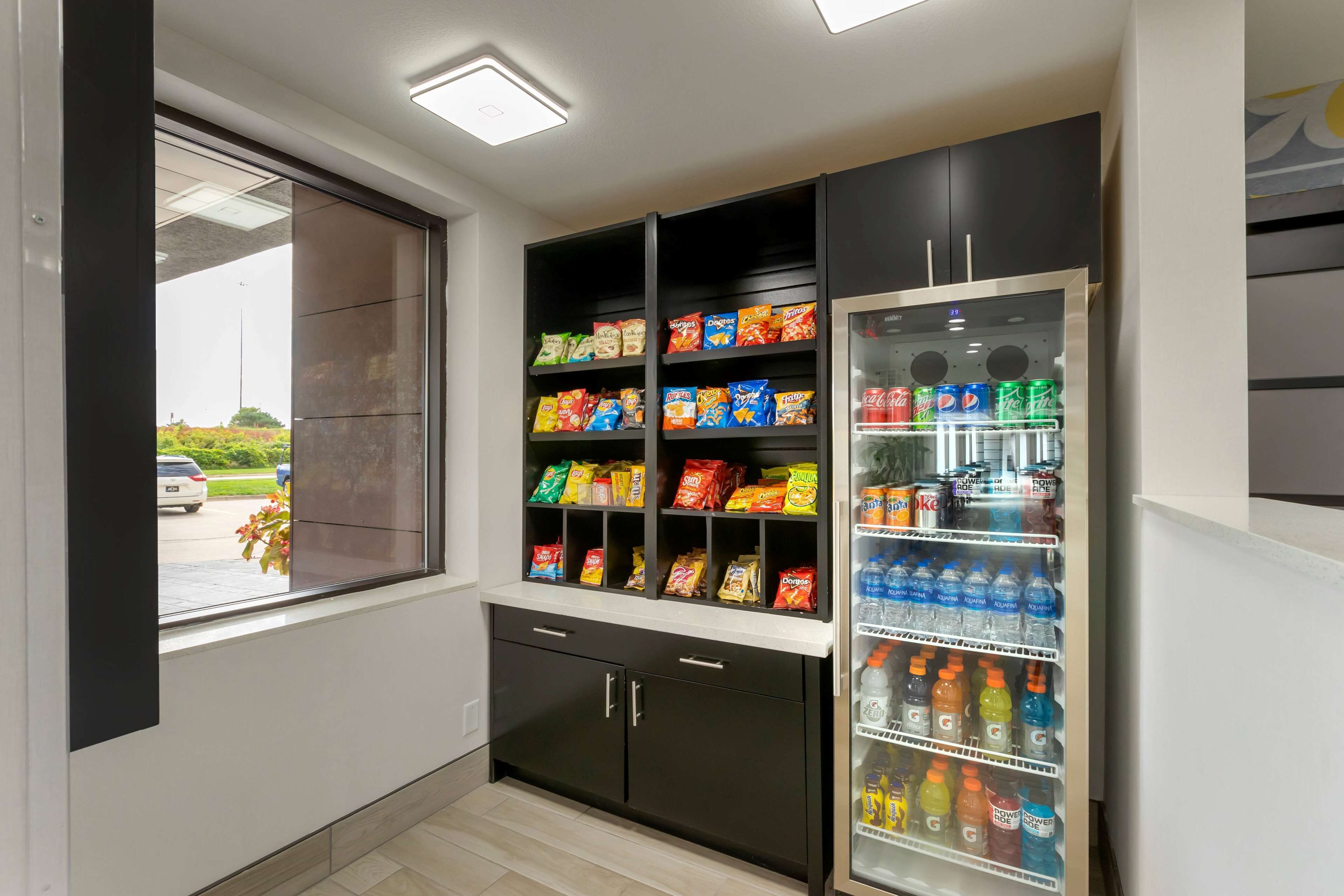 A kitchenette with a refrigerator stocked with beverages and snacks, a window with a view of a parking lot, and a counter with a sink.