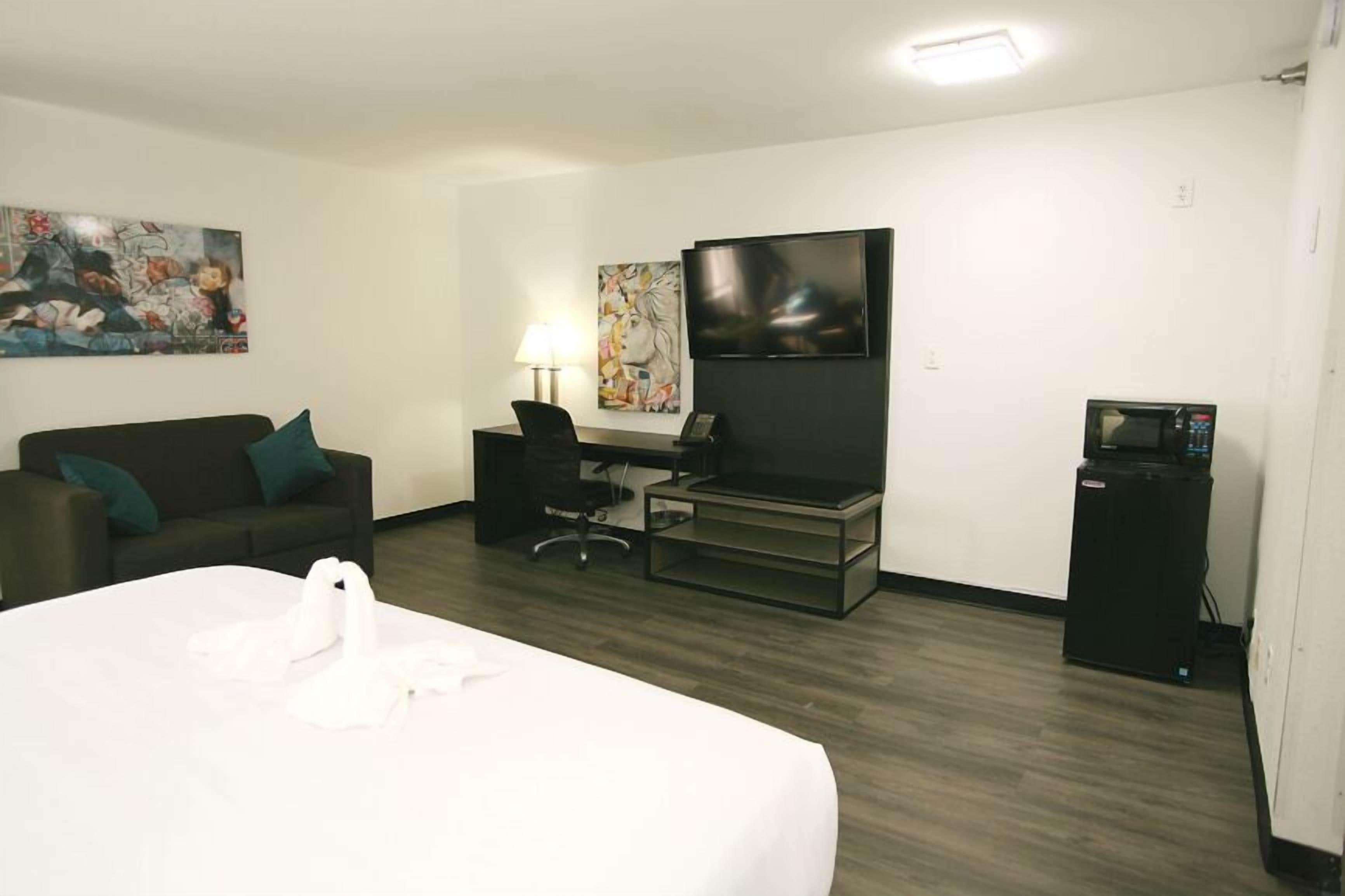 A hotel room with a bed, a desk with a chair, a TV, a microwave, and artwork on the walls.