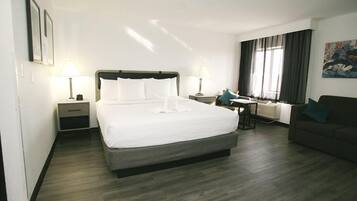 A hotel room with a large bed, two bedside tables with lamps, a small table with a chair, a sofa, and a large window with curtains.