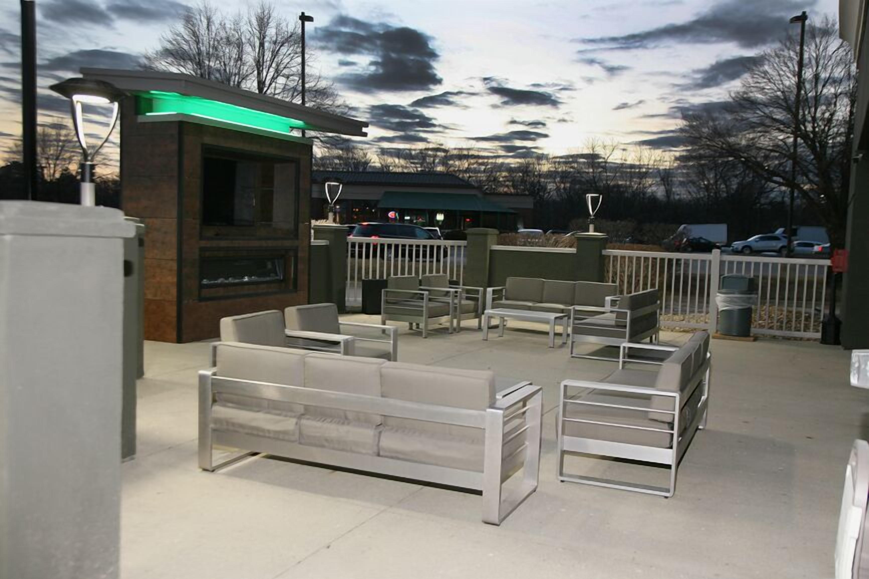 An outdoor seating area with modern furniture, a building with a green-lit entrance, and a parking lot in the background.