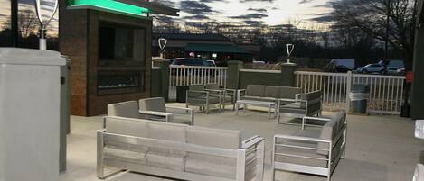 Un área de estar al aire libre con mobiliario moderno, un edificio con entrada iluminada en verde, y un estacionamiento al fondo.