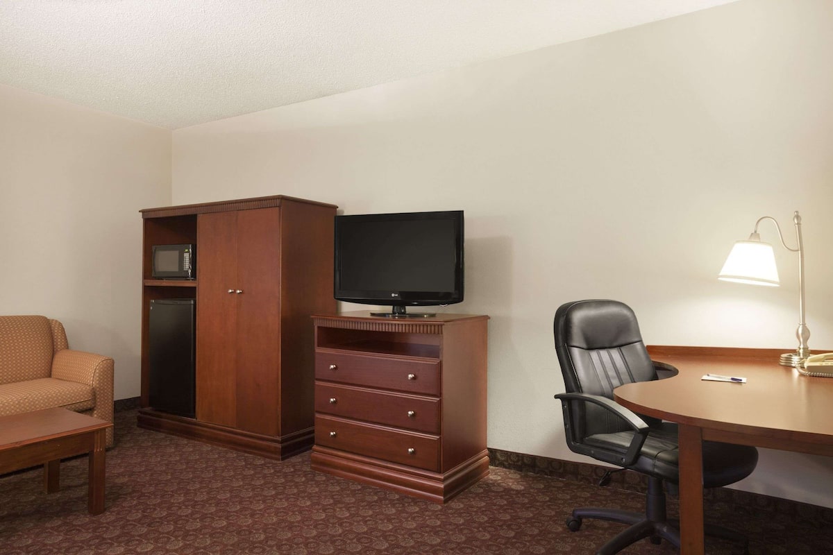 Ein Hotelzimmer mit einem Holzkasten, einem Fernseher, einem Schreibtisch mit Lampe, einem Sessel und einem gemusterten Teppich.