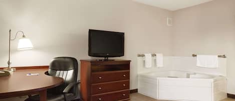 A hotel room with a desk, chair, television, and a large bathtub.