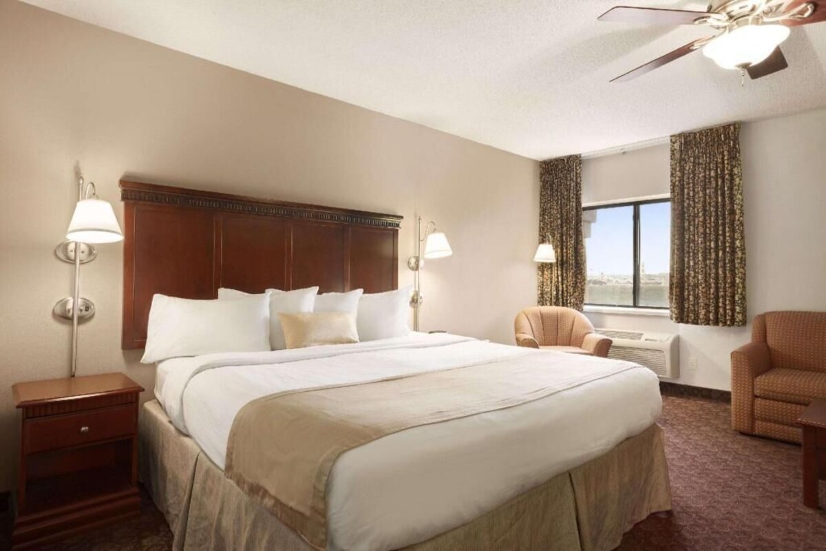 A hotel room with a large bed, two bedside lamps, a ceiling fan, a window with curtains, and a chair.