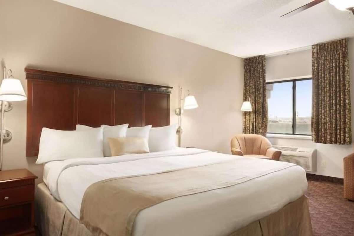 Ein Hotelzimmer mit einem großen Bett, zwei Nachttischlampen, einem Sessel und einem mit Vorhängen versehenen Fenster.