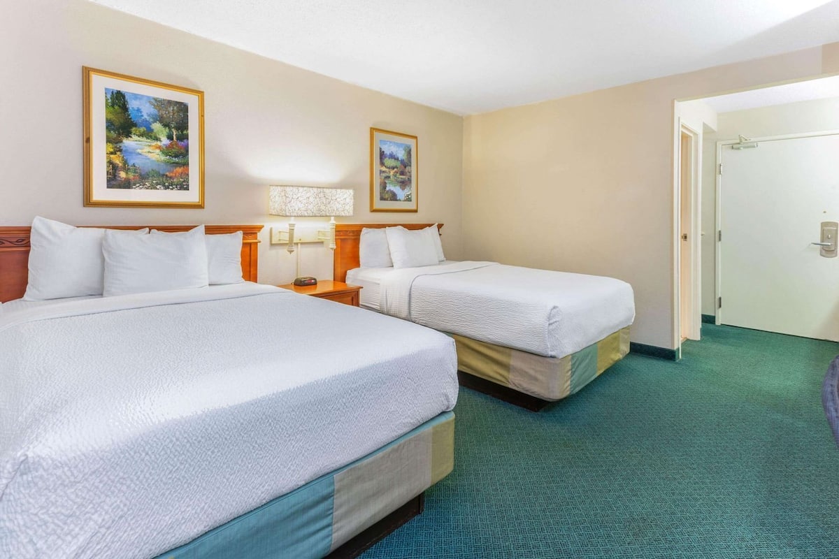 A hotel room with two beds, a nightstand with a lamp, and two framed paintings on the wall.
