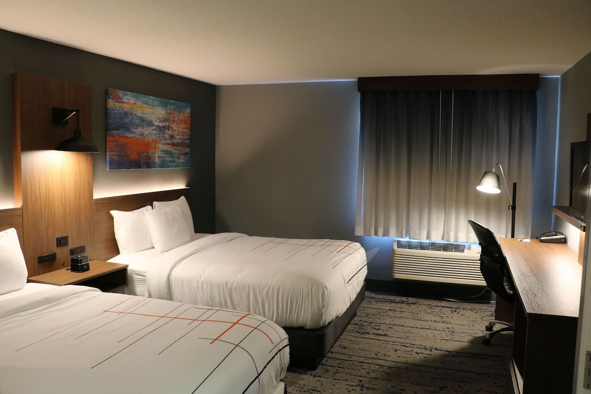 A hotel room with two beds, a desk, a chair, a desk lamp, a painting, and an air conditioning unit.