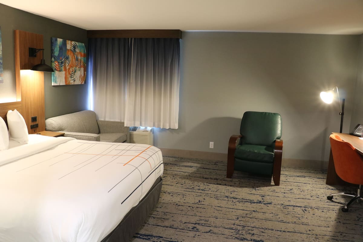 A hotel room with a large bed, a green armchair, a desk with a lamp, and a painting on the wall.