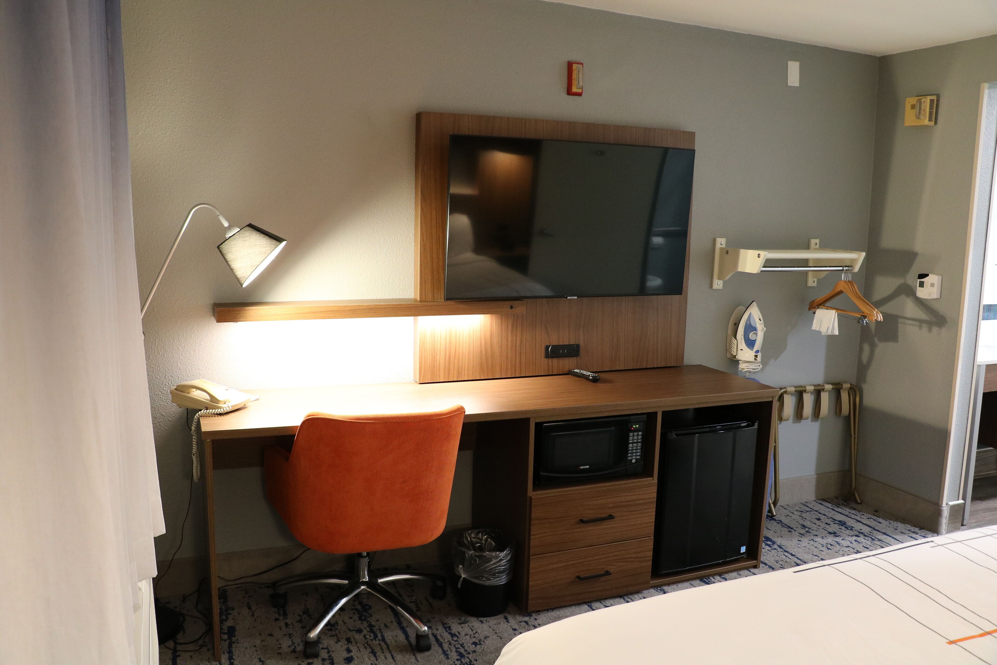 Une chambre d’hôtel équipée d’un bureau en bois, d’une chaise orange, d’un téléviseur à écran plat, d’un four à micro-ondes, d’un mini-réfrigérateur, d’une poubelle et d’un porte-vêtements.