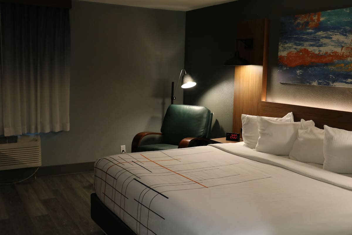 A hotel room with a large bed, a green chair, a desk with a lamp, and a painting on the wall.