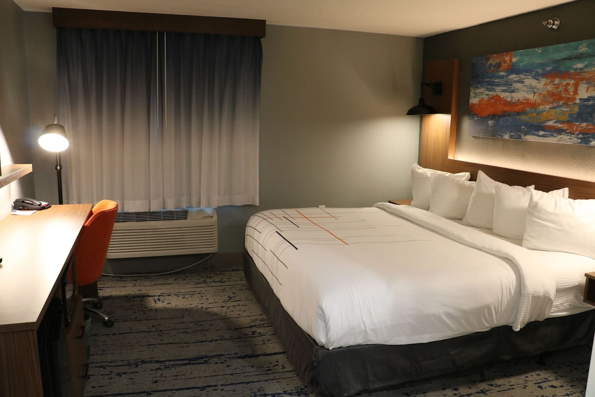 A hotel room with a bed, a desk with a lamp, a chair, and a painting on the wall.