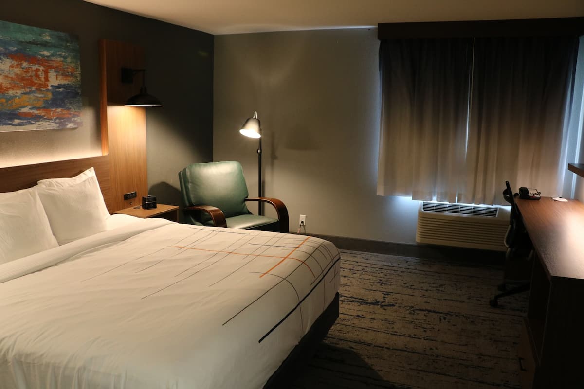 A hotel room with a large bed, a desk, a chair, a lamp, a painting on the wall, and a window with curtains.