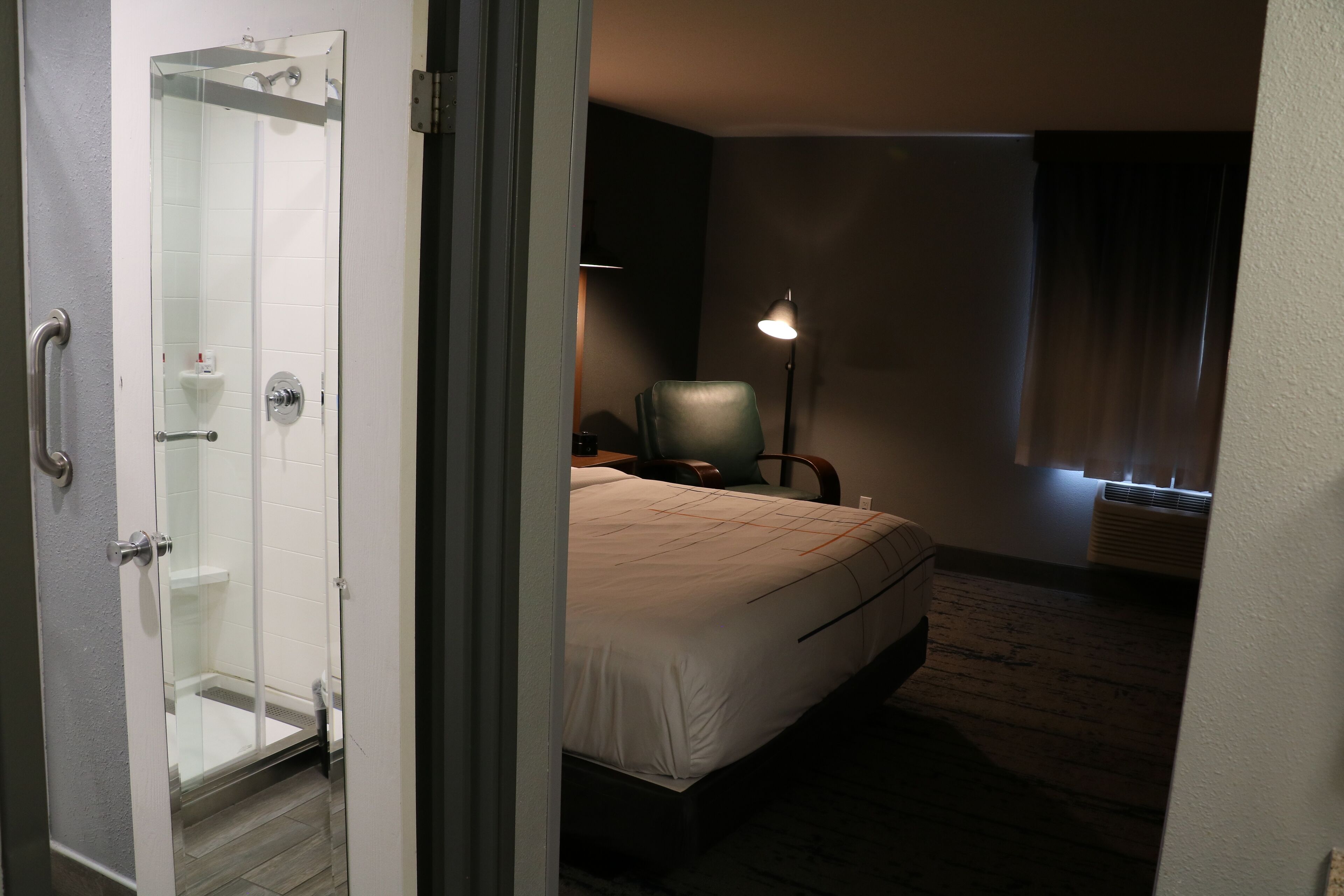 A hotel room with a bed, a chair, a desk, a lamp, and a shower area.