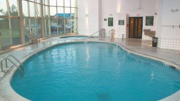 Una piscina interior con paredes de vidrio transparente, suelo de baldosas y algunas sillas alrededor.