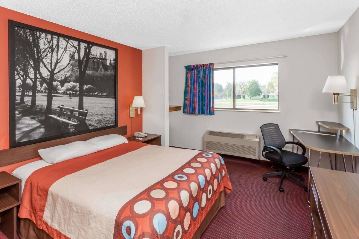 Habitación de hotel con cama, escritorio, silla, ventana y un cuadro grande enmarcado en la pared.