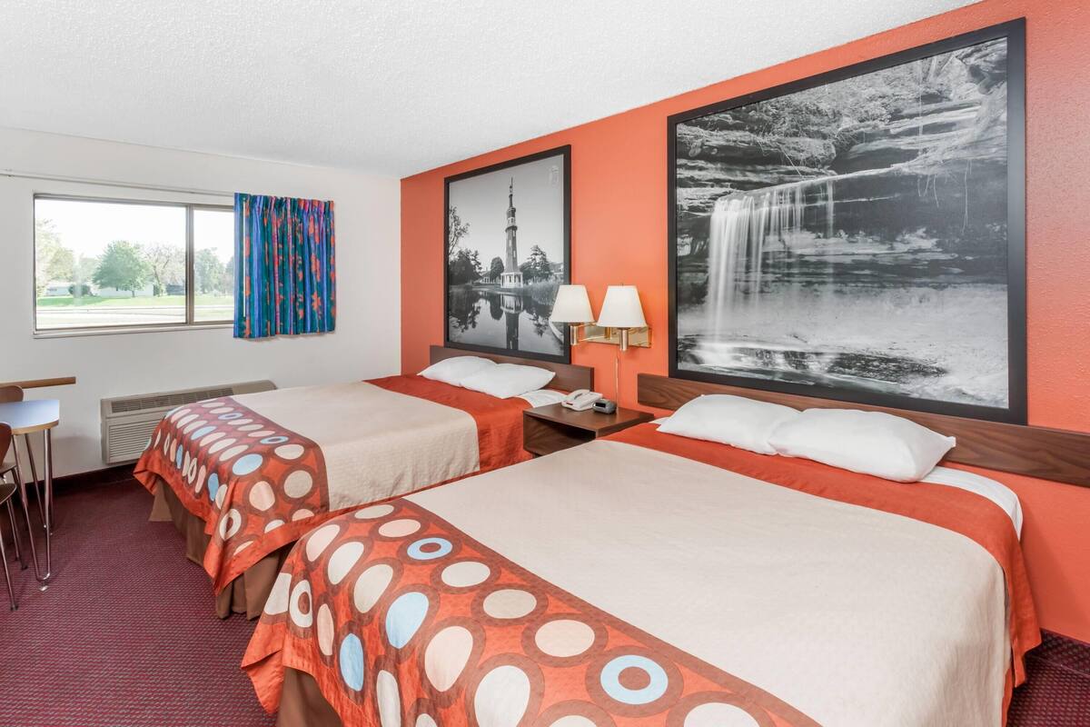 Una habitación de hotel con dos camas, una ventana con cortinas, una lámpara de pared y dos cuadros enmarcados.