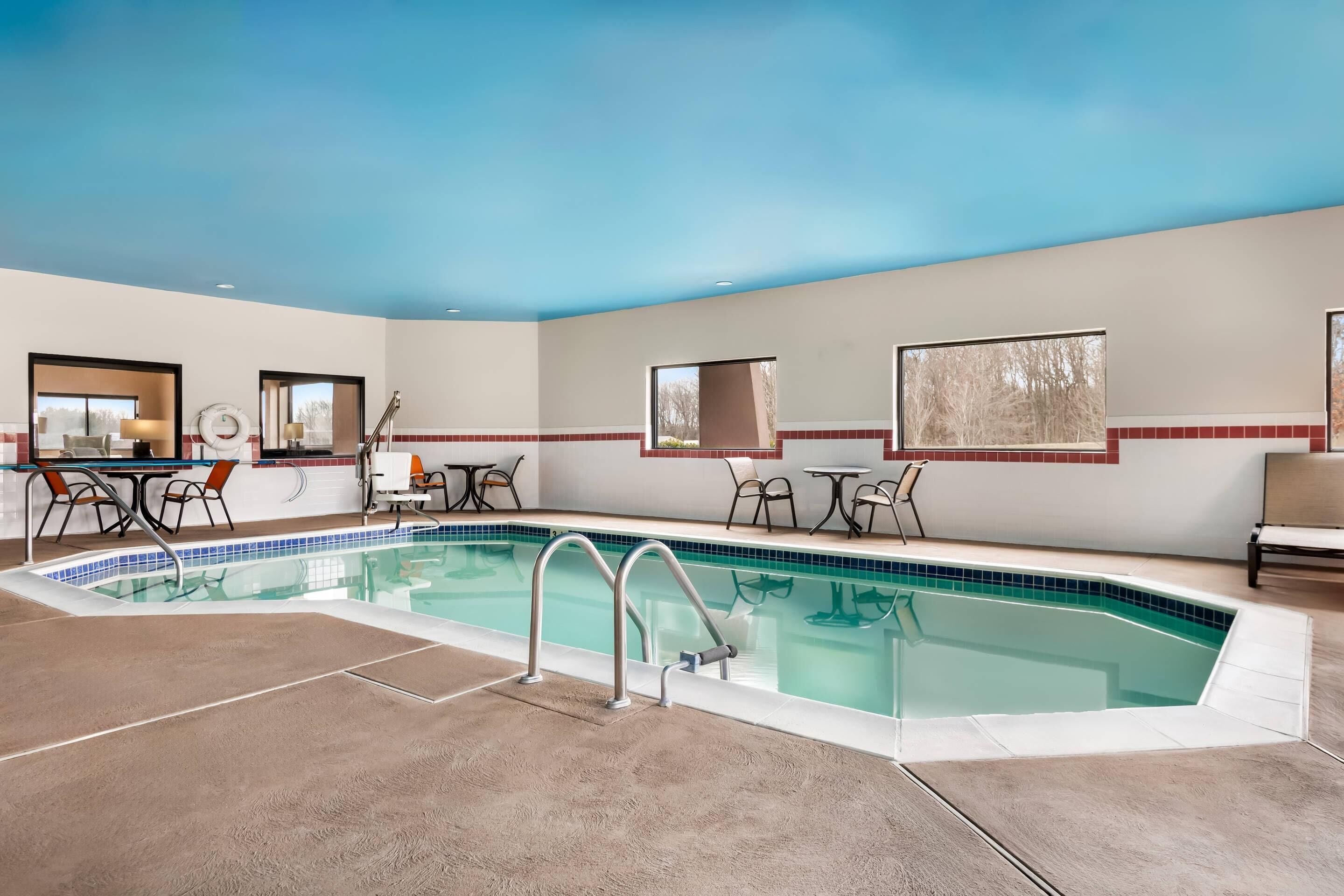 Una piscina interior con borde de azulejos, rodeada por un suelo y paredes beige con detalles rojos. Hay mesas y sillas alrededor de la piscina, y una puerta que conduce a otra habitación.