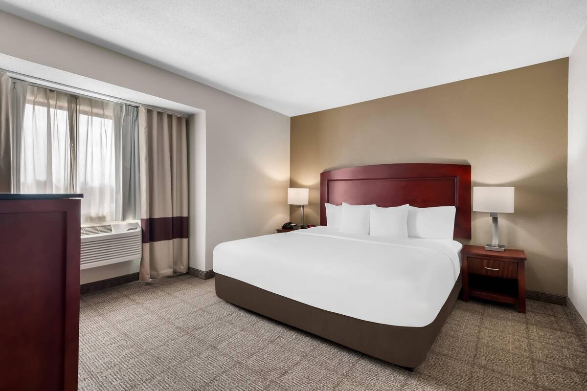 A hotel room with a large bed, a bedside table with a lamp, a nightstand, a window with curtains, and a radiator.