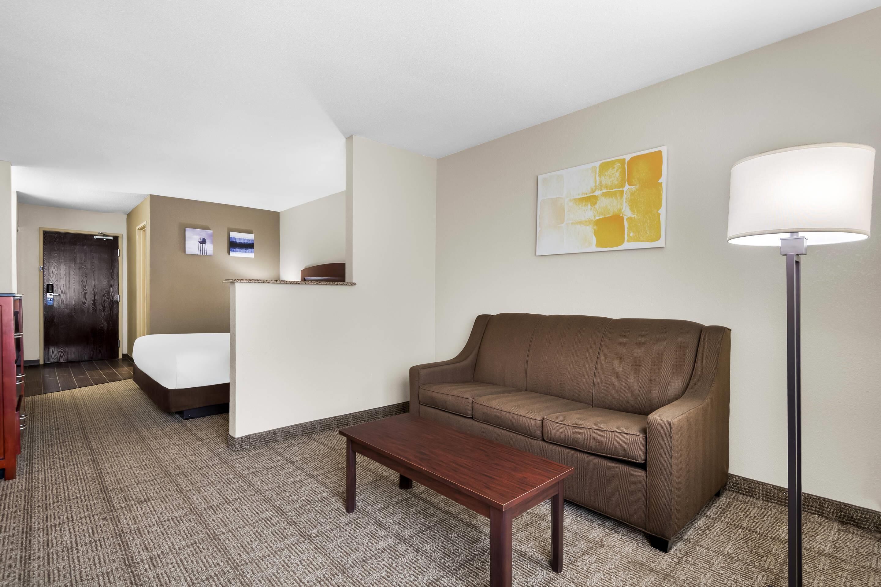 Una habitación de hotel con un sofá marrón, una mesa de centro de madera, una lámpara y una cama.