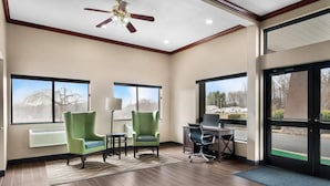 Une chambre d’hôtel avec deux fauteuils verts, un bureau avec un ordinateur portable, un ventilateur de plafond et de grandes fenêtres donnant sur des arbres et une route.
