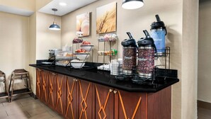 Un buffet de petit-déjeuner d’hôtel avec des distributeurs de café et d’eau, un saladier de fruits et un panier de pain.