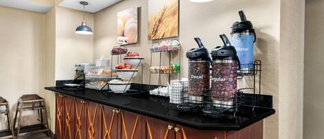 A hotel breakfast bar with coffee and water dispensers, a fruit bowl, and a bread basket.