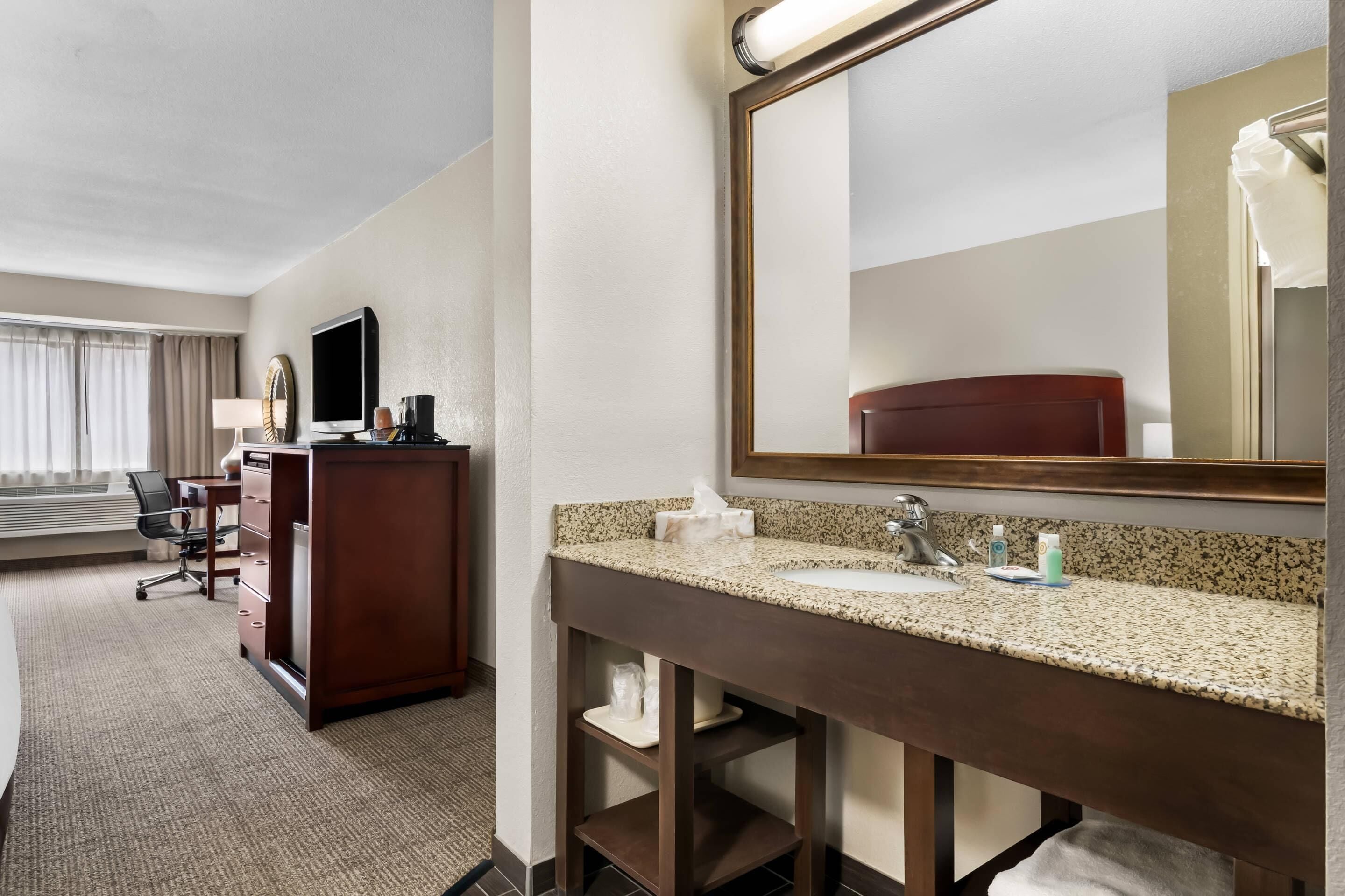 A hotel bathroom with a large mirror, a sink, a vanity, a desk, and a chair.
