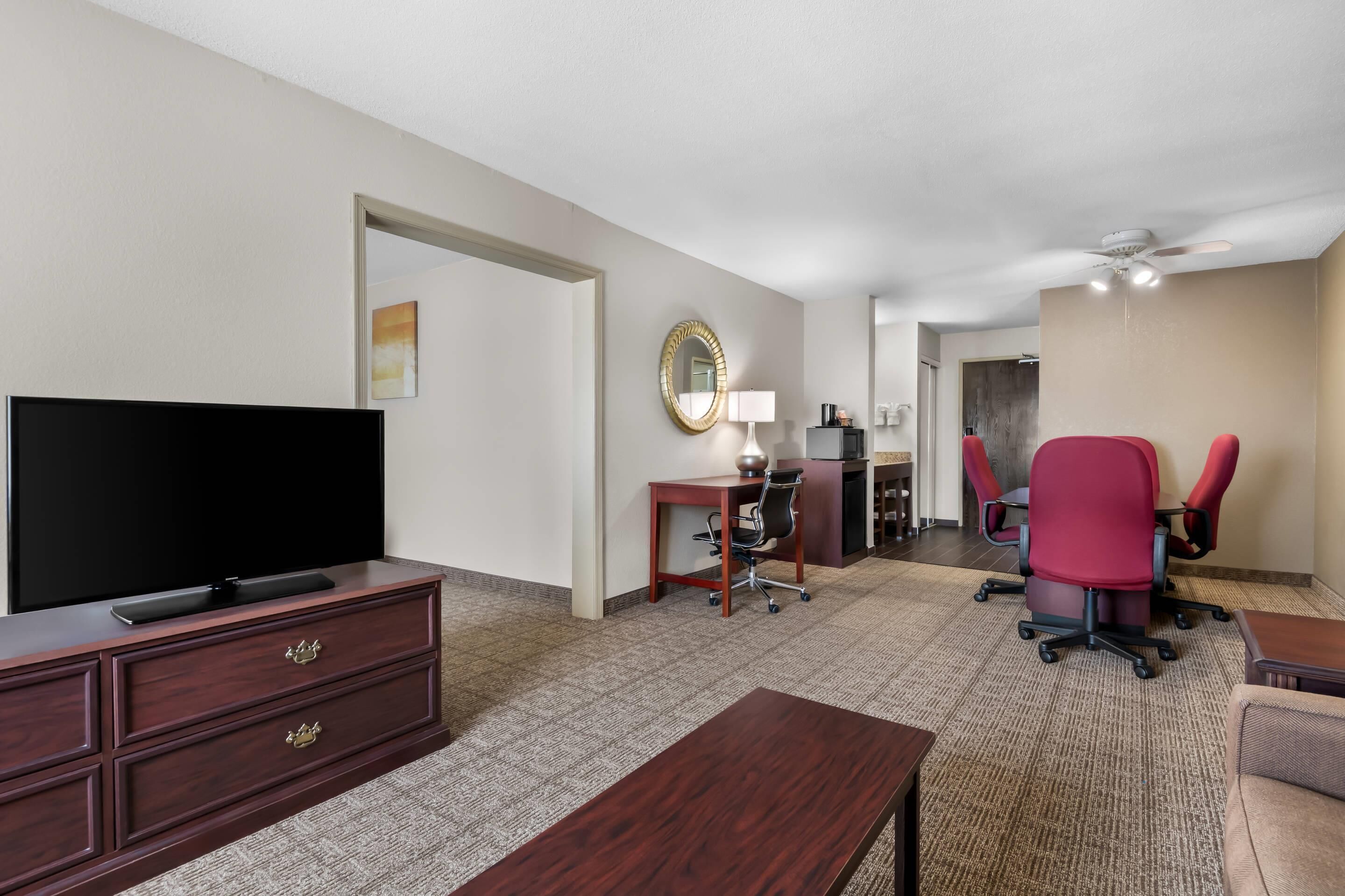 Ein Hotelzimmer mit Flachbildfernseher, einem Holzkoffer, einem Schreibtisch mit Stuhl, einem roten Bürostuhl und einem Deckenventilator.