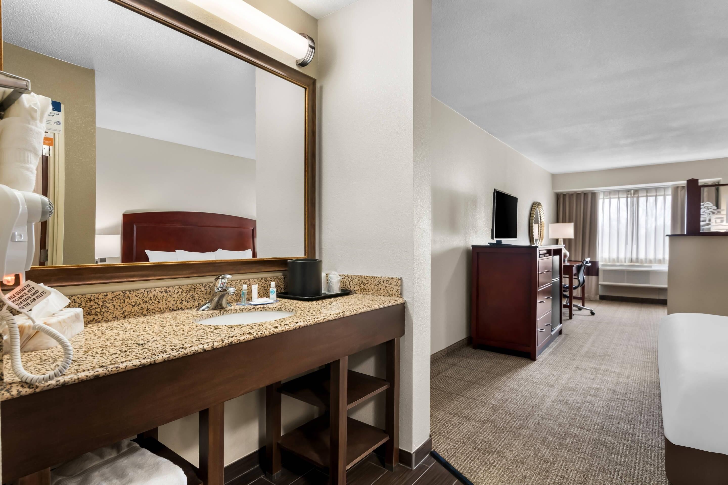 A hotel bathroom with a granite countertop sink, a mirror, a hairdryer, and a robe rack.