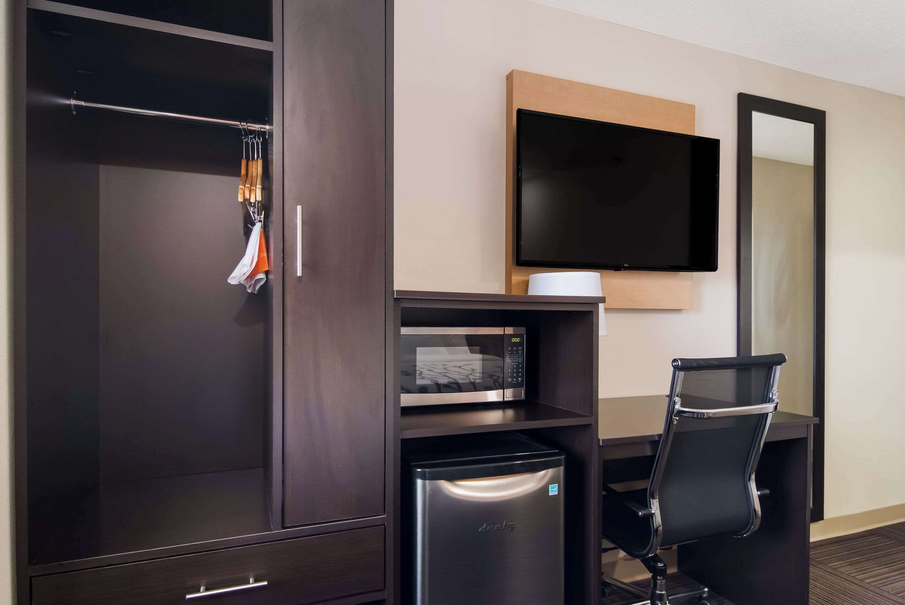 A hotel room with a dark wooden wardrobe, a small refrigerator, a desk with a chair, a flat-screen TV, and a mirror.