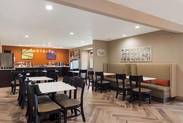 A modern dining area with tables and chairs, a kitchen in the background, and a seating area with a sofa and cushions.