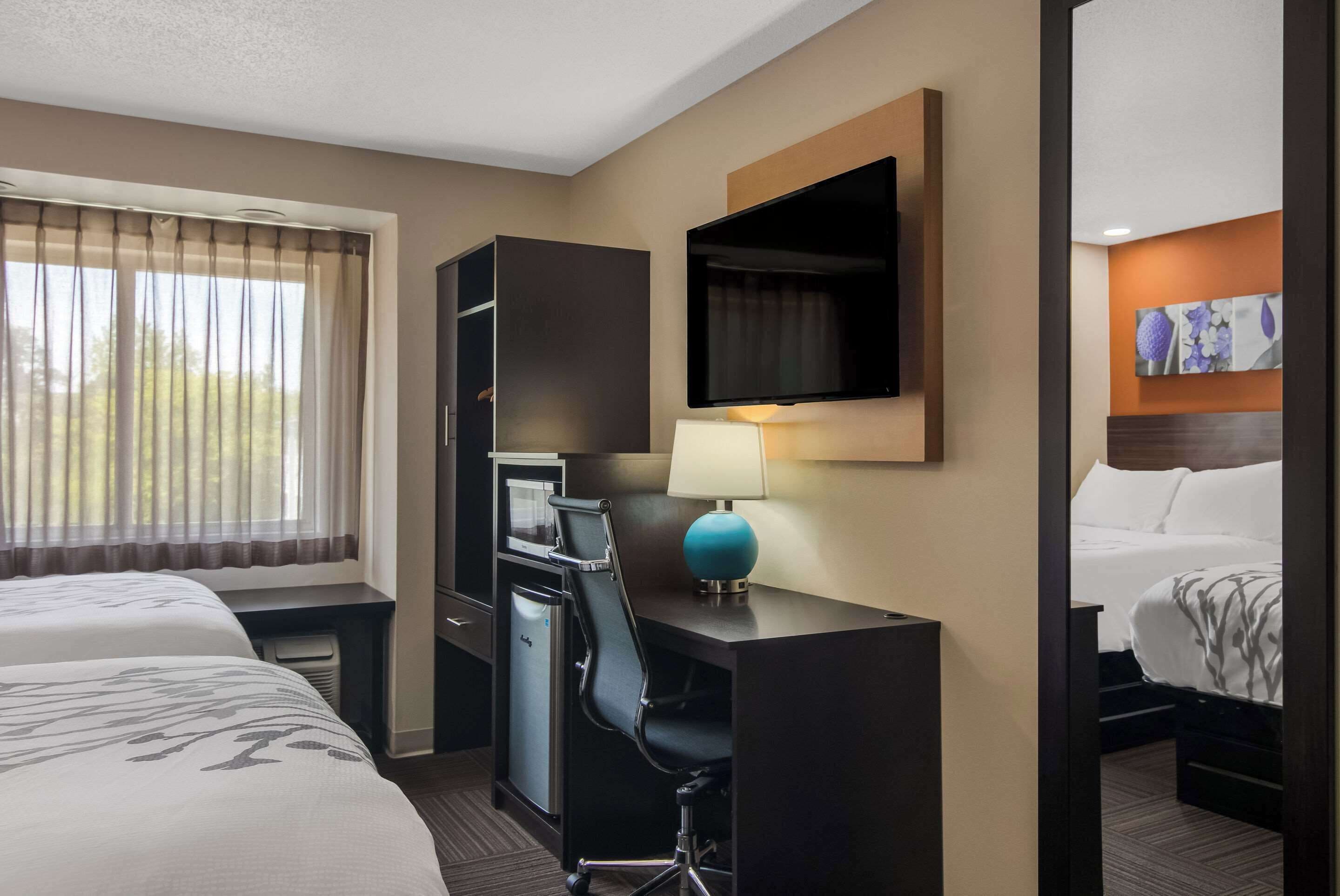 A hotel room with two beds, a desk with a lamp, a television, and a small kitchenette.