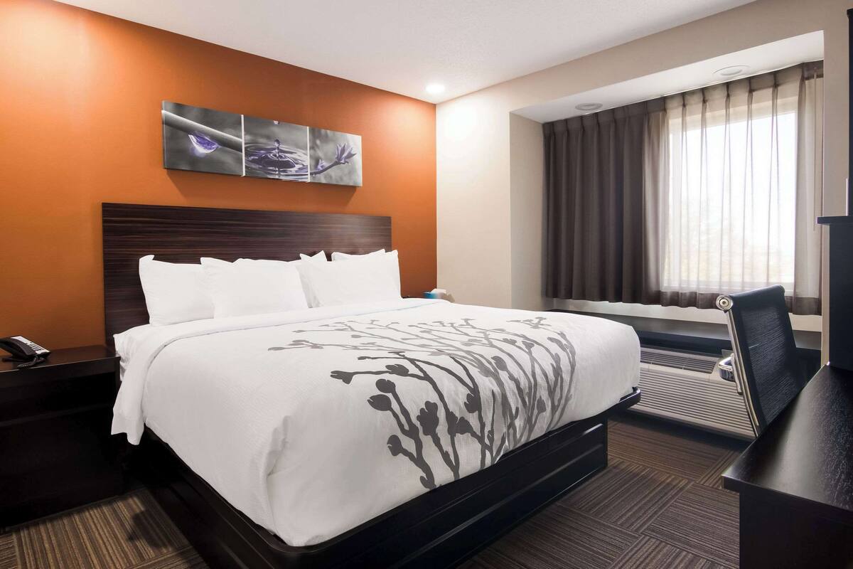 A hotel room with a large bed, a nightstand with a phone, a desk with a chair, and a window with curtains.