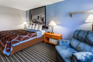 Habitación de hotel con cama, dos lámparas de noche, mesita de noche con radio, un sillón azul y un cuadro de un paisaje urbano en la pared.
