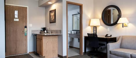 A hotel room with a wooden door, a kitchenette, a desk with a chair, a round mirror, and a sofa.