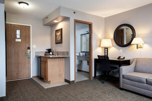 Hotellihuone, jossa on puinen ovi, pieni keittiönurkkaus, työpöytä tuolilla, pyöreä peili ja sohva.