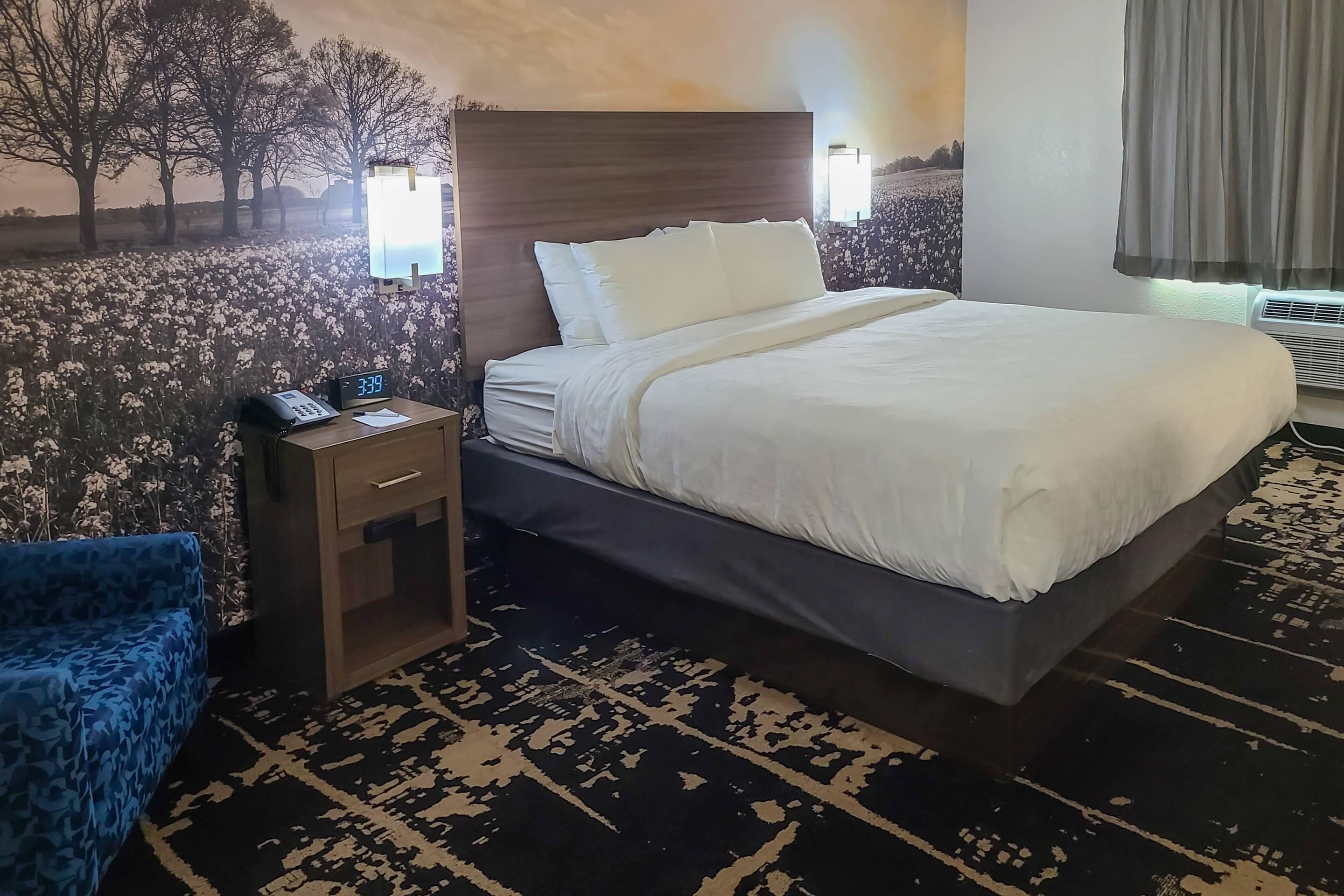 A hotel room with a large bed, a bedside table with a phone, a blue chair, and a patterned rug.